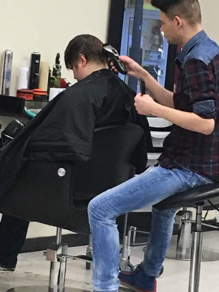 Strzyżenie włosów maszynką w salonie: klient w czarnej pelerynie siedzi na fotelu, stylista w dżinsowej koszuli i jeansach używa maszynki na włosach klienta.