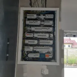 Otwarta skrzynka rozdzielcza z zainstalowanymi bezpiecznikami i okablowaniem w surowym wnętrzu, widoczne przewody w ścianie i gniazdko elektryczne poniżej, na zewnątrz budynku widok na dom...