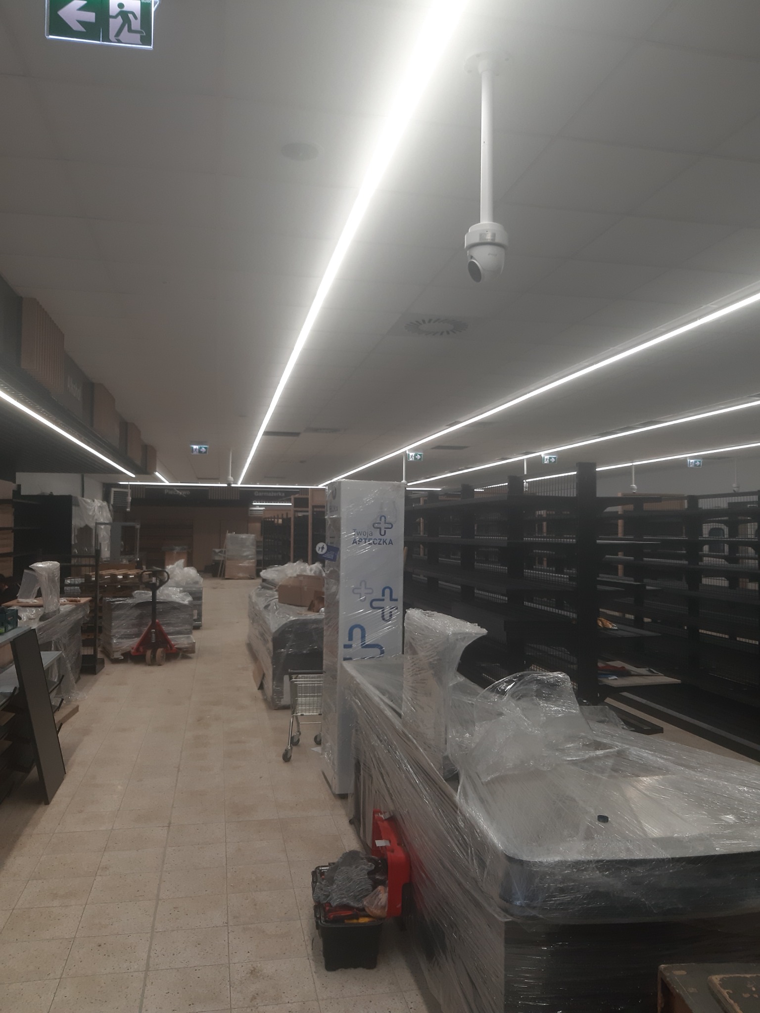Wnętrze sklepu w trakcie aranżacji: widoczne regały, oświetlenie LED, kamera monitoringu sufitowa, wózki transportowe i elementy wyposażenia zabezpieczone folią stretch.