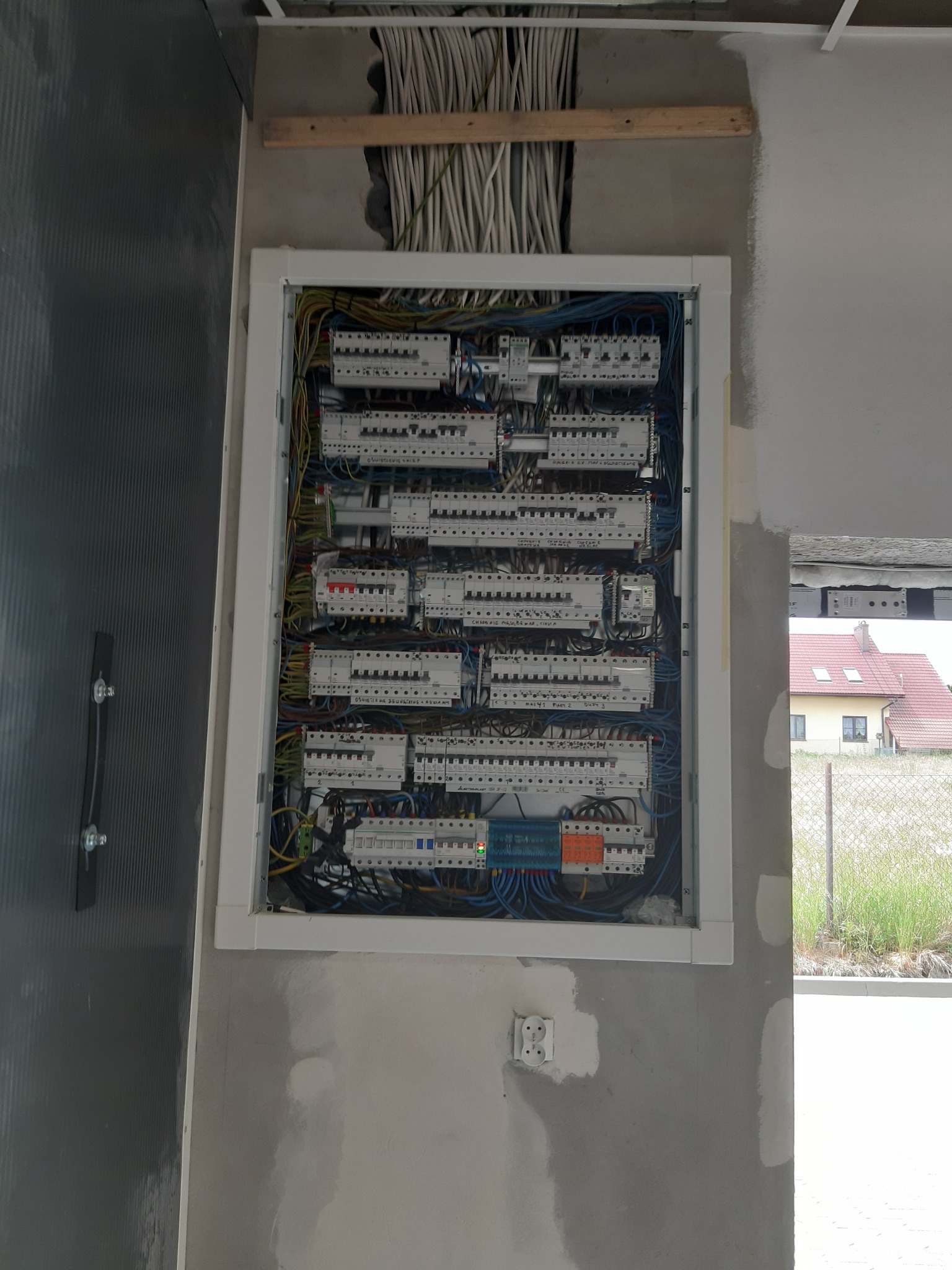 Otwarta skrzynka rozdzielcza z zainstalowanymi bezpiecznikami i okablowaniem w surowym wnętrzu, widoczne przewody w ścianie i gniazdko elektryczne poniżej, na zewnątrz budynku widok na dom...