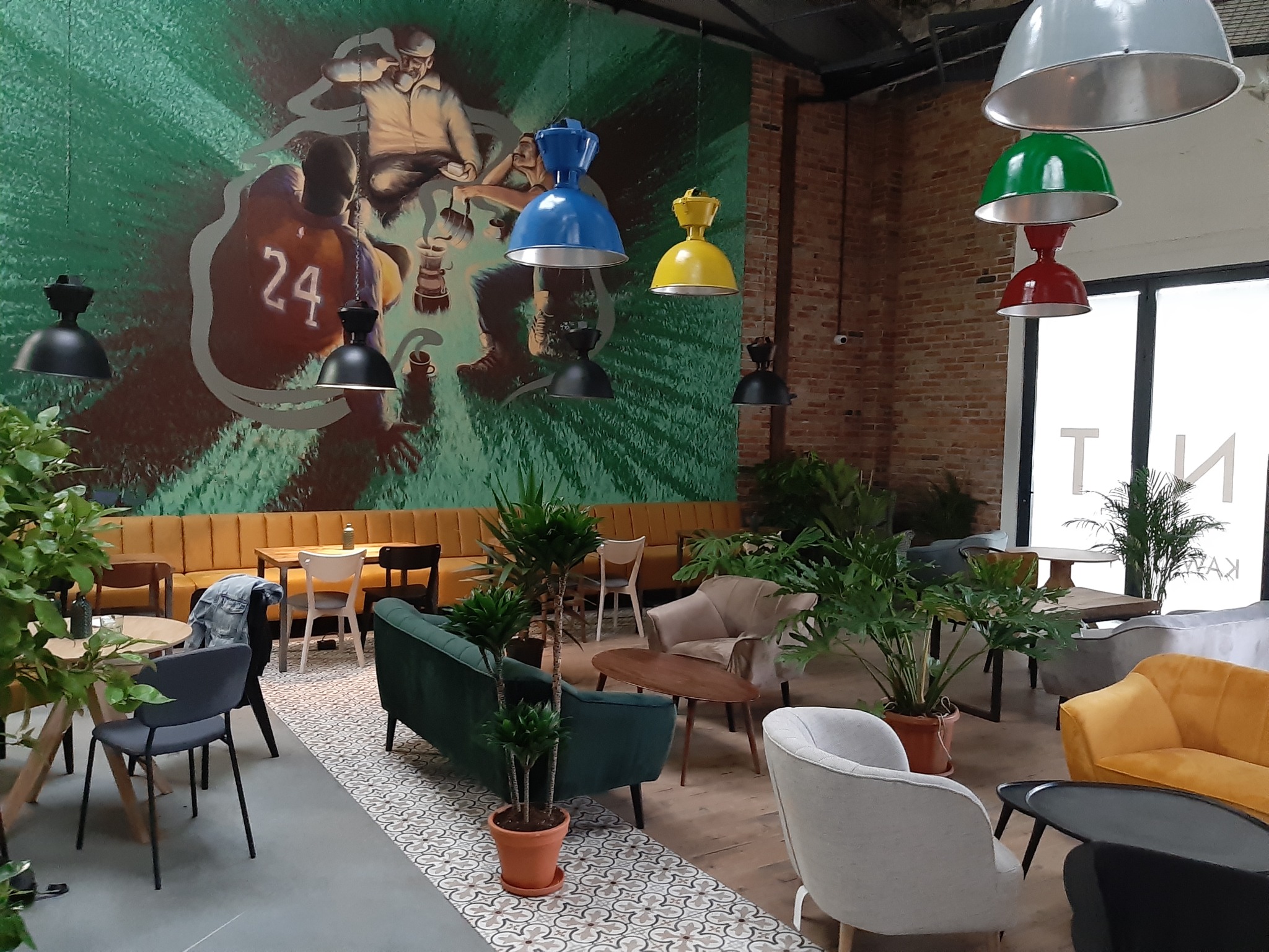 Wnętrze kawiarni z muralem przedstawiającym koszykarza z numerem 24 i starszego mężczyznę, wiszące kolorowe lampy w stylu przemysłowym, rośliny doniczkowe i różnorodne meble wypoczynkowe.
