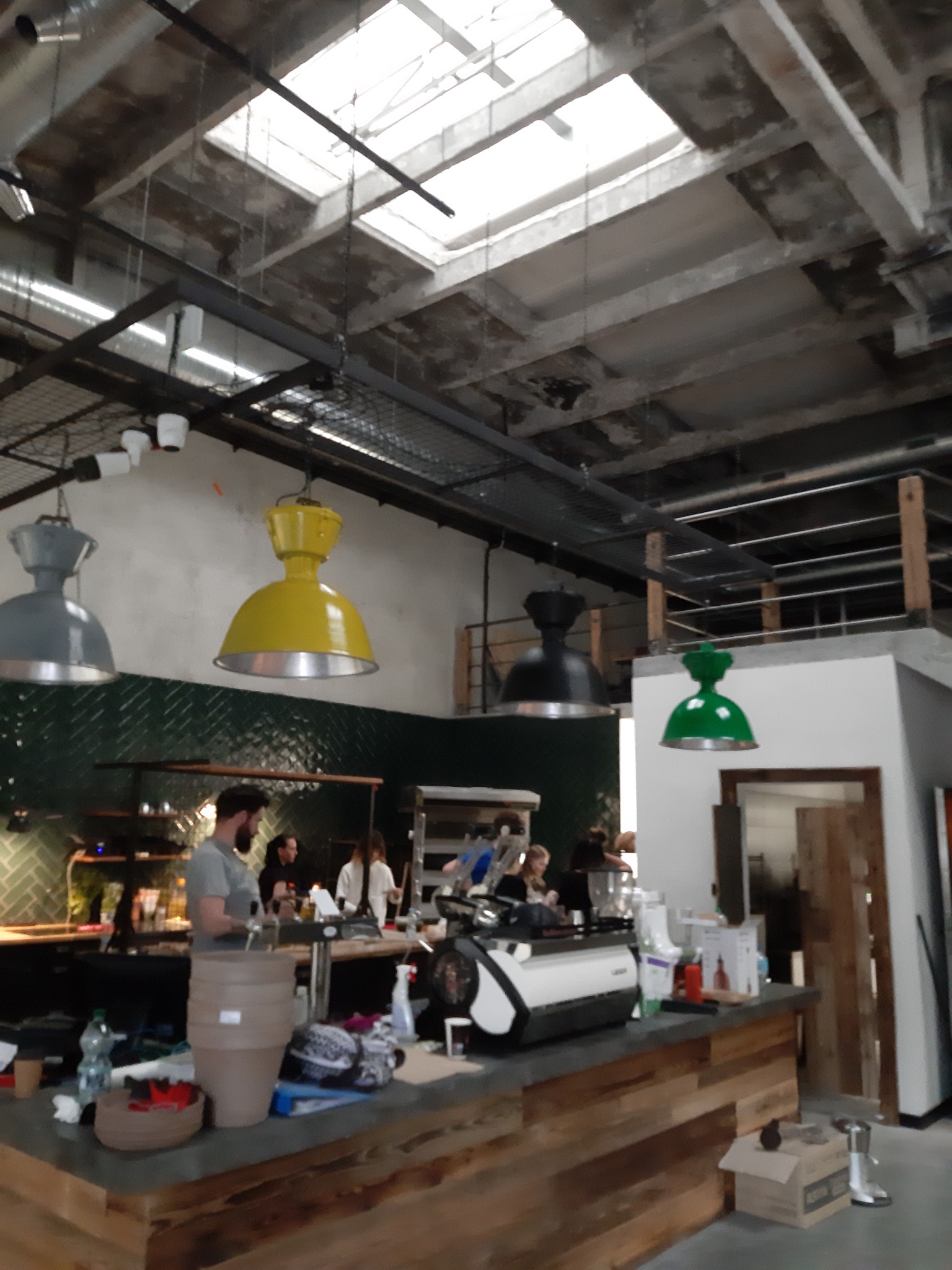 Wnętrze lokalu gastronomicznego z widocznymi lampami w kolorach szarym, żółtym, czarnym i zielonym, zamontowanymi pod betonowym sufitem z oknami dachowymi, oraz z barem z ekspresem do kawy...