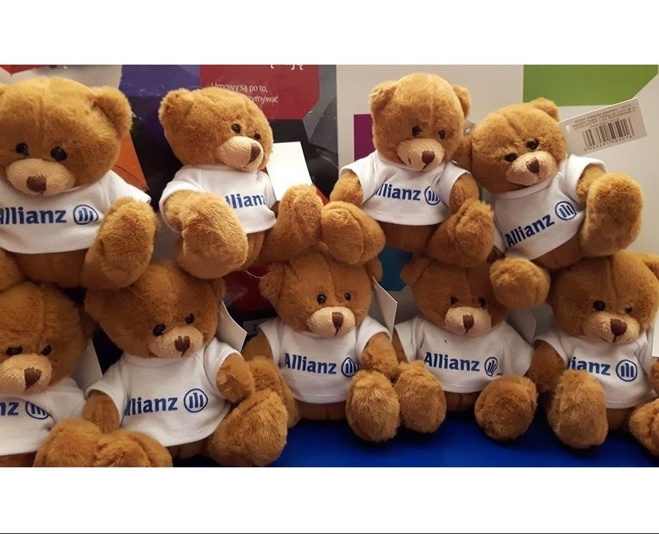 Grupka pluszowych misiów w białych koszulkach z logo firmy ubezpieczeniowej, ustawionych blisko siebie na niebieskim tle.