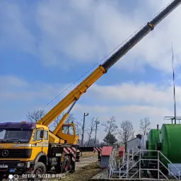 Żółty dźwig samochodowy Mercedes-Benz z wysuniętym ramieniem na tle błękitnego nieba z chmurami, obok instalacji przemysłowej z zielonymi zbiornikami.