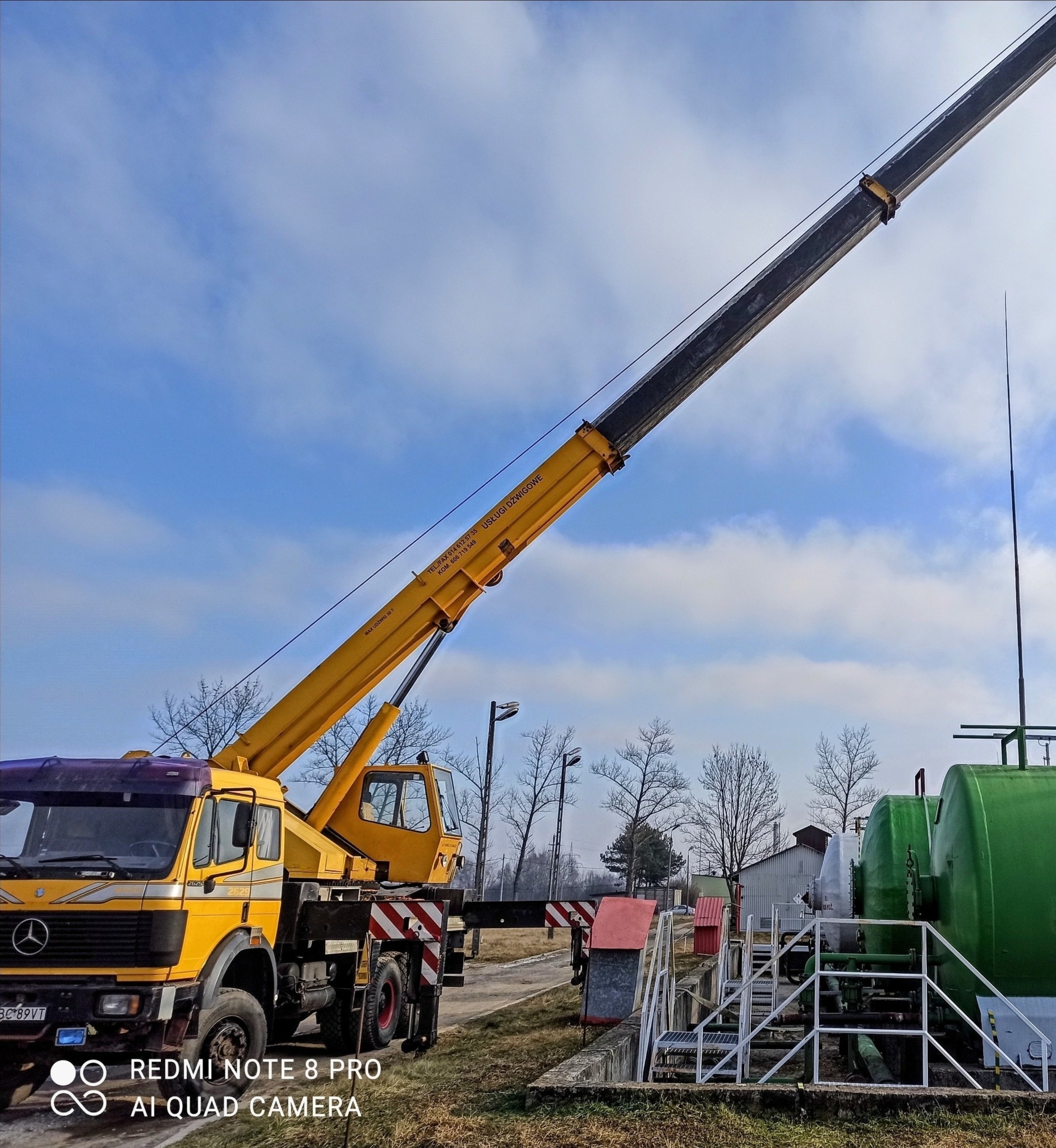 Żółty dźwig samochodowy Mercedes-Benz z wysuniętym ramieniem na tle błękitnego nieba z chmurami, obok instalacji przemysłowej z zielonymi zbiornikami.