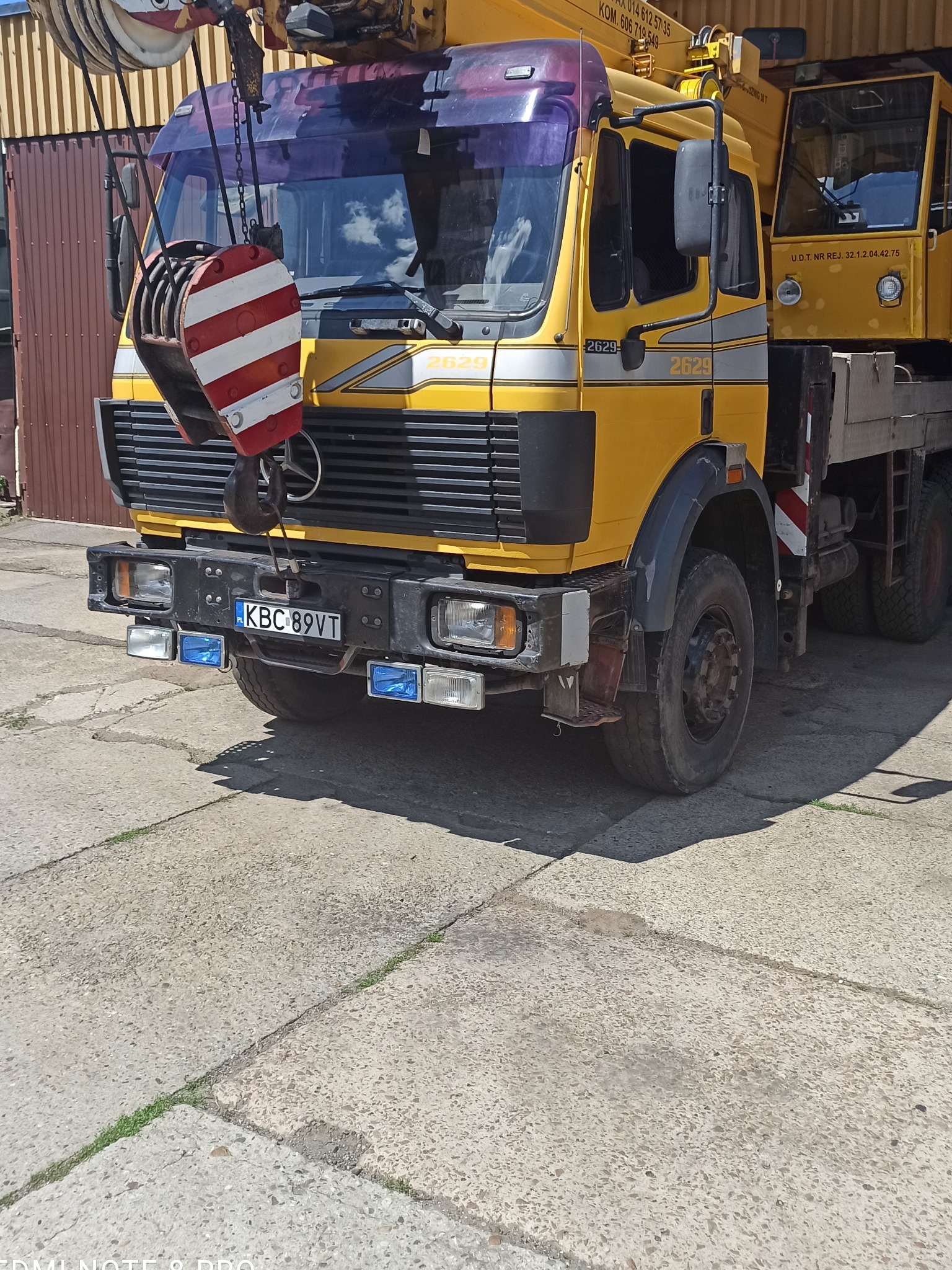Żółty samochód ciężarowy z dźwigiem i hakiem w biało-czerwone pasy, zaparkowany na betonowym placu, z widocznymi numerami rejestracyjnymi i oznaczeniami UDT.