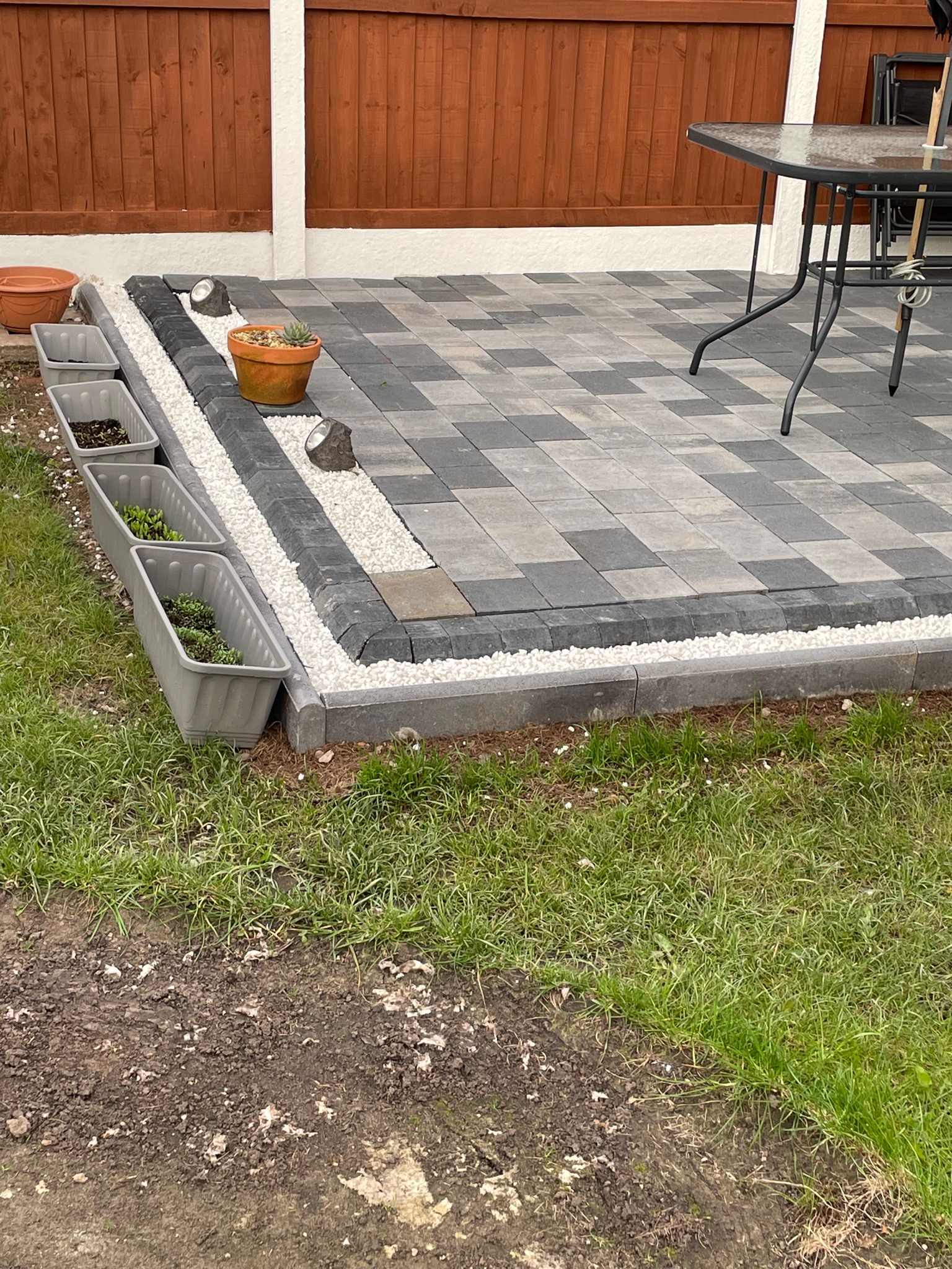 Nowoczesny taras z kostki brukowej w szaro-czarną szachownicę, obrzeża z białego grysu, donice z sadzonkami i stolik ogrodowy w tle drewnianego ogrodzenia.