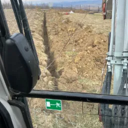 Wąski wykop w ziemi widziany z kabiny koparki, z widocznymi elementami sterowania i naklejkami informacyjnymi na szybie, w tle krajobraz z polami i zabudowaniami.