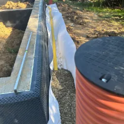 Wykop pod budynek z zabezpieczonymi ścianami i izolacją przeciwwilgociową, widoczny poziomica, rura drenarska, geowłóknina i studzienka kanalizacyjna.