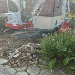 Mała koparka Takeuchi TB225 na placu budowy, przygotowująca teren pod fundamenty budynku z czerwonej cegły, widoczne fragmenty starego chodnika i roślinność.
