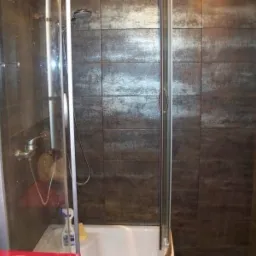 Kabina prysznicowa z przesuwnymi drzwiami, wykończona ciemnymi, metalizowanymi płytkami w kwadratowym formacie, widoczna bateria i słuchawka prysznicowa.