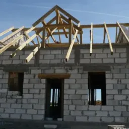 Budowa domu jednorodzinnego: ściany z bloczków betonowych, drewniana konstrukcja dachu w trakcie montażu, widoczne otwory okienne i drzwiowe.