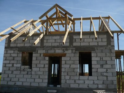 Budowa domu jednorodzinnego: ściany z bloczków betonowych, drewniana konstrukcja dachu w trakcie montażu, widoczne otwory okienne i drzwiowe.