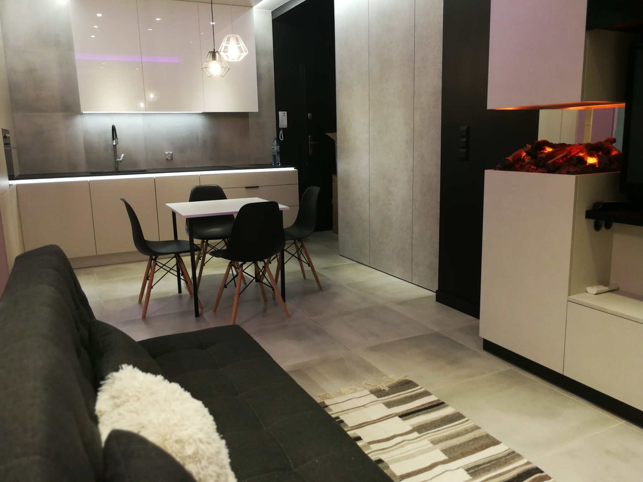 Nowoczesne wnętrze salonu z aneksem kuchennym we Wrocławiu, z szarą sofą z dekoracyjną poduszką, stołem z krzesłami i imitacją kominka w zabudowie.