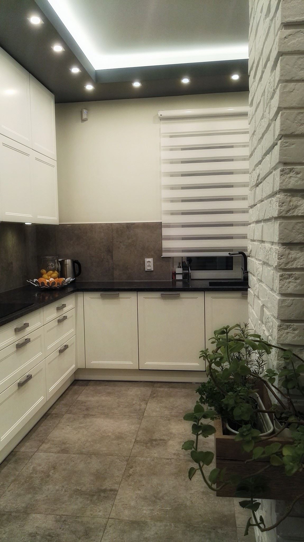 Jasna kuchnia z białymi szafkami, czarnym blatem, szarą ścianą nad blatem i ceglaną ścianą, roletą okienną oraz roślinami w doniczce.