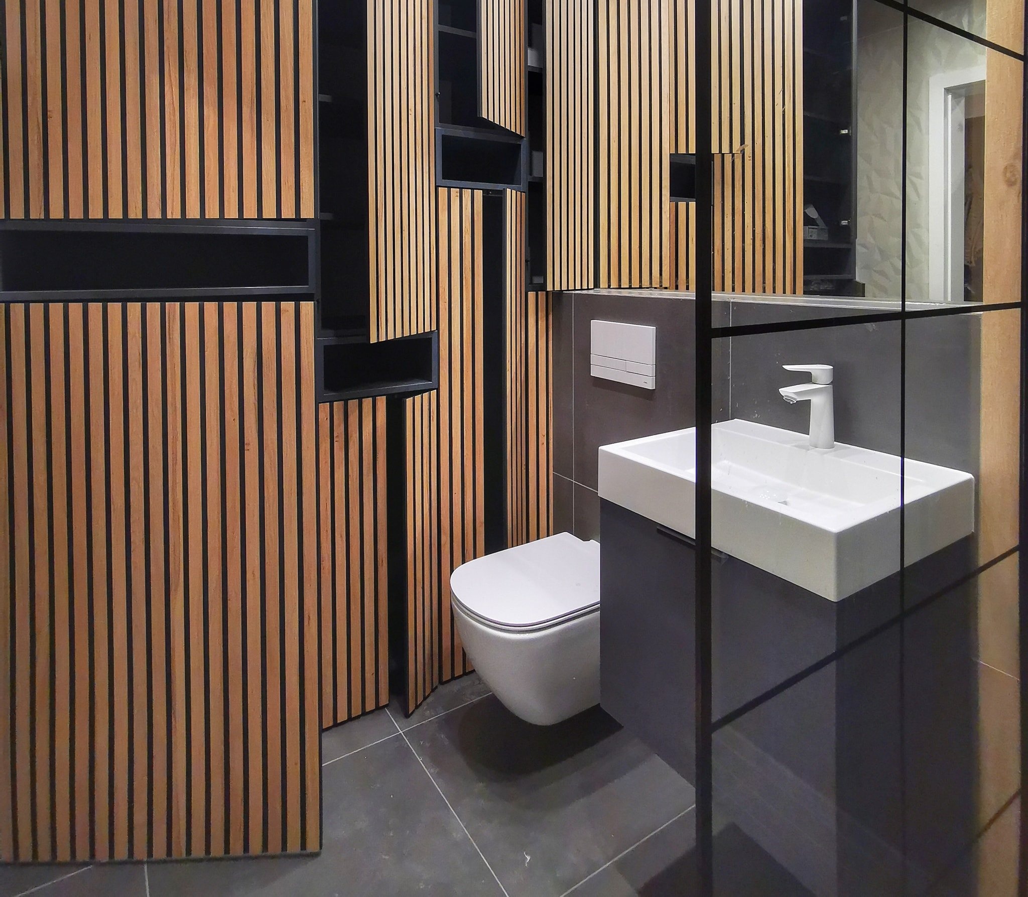 Nowoczesna łazienka z drewnianymi lamelami na ścianie, wisząca toaleta i umywalka nablatowa z grafitową szafką.