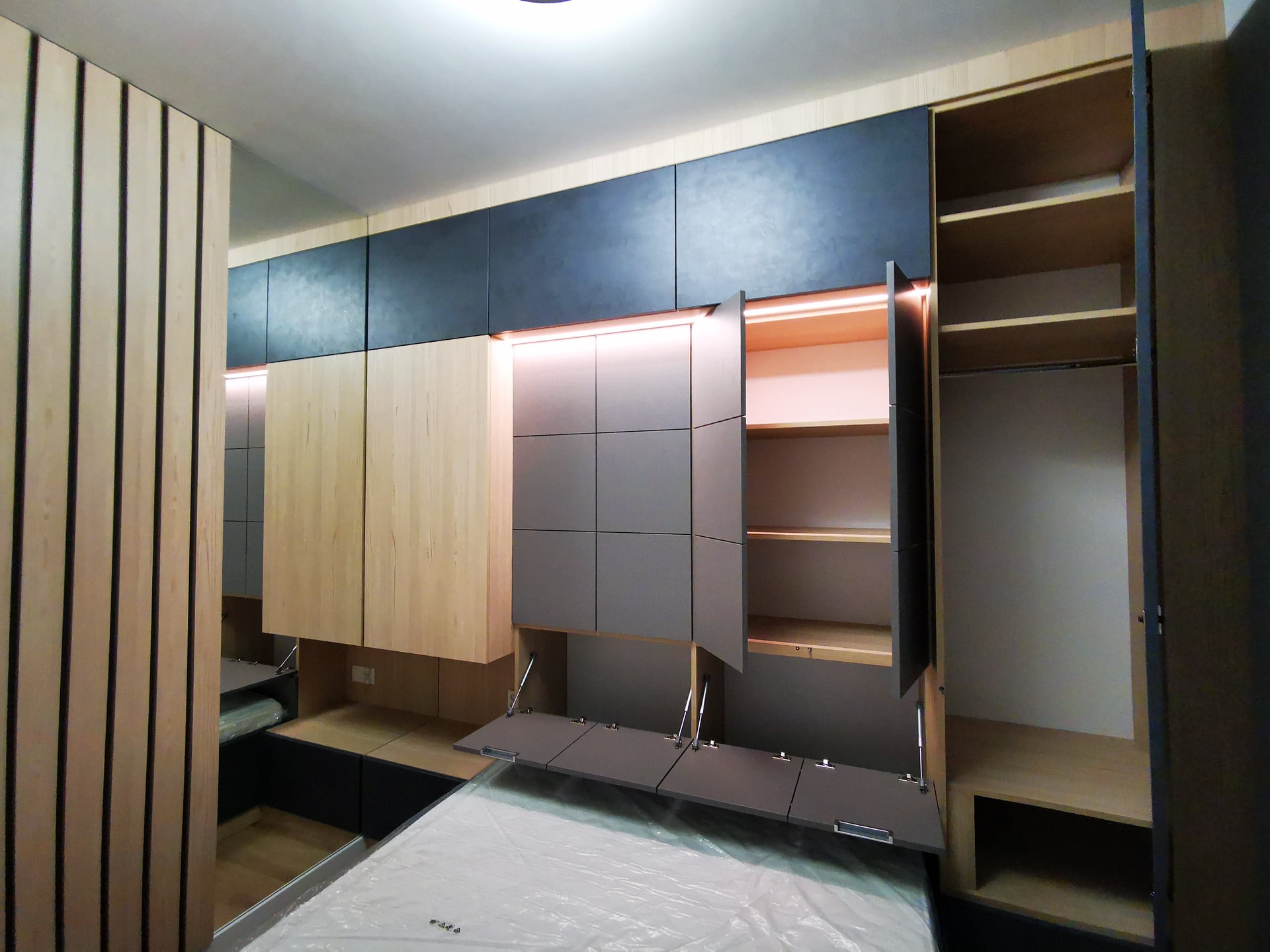 Nowoczesna sypialnia z zabudową meblową: szafy z frontami w odcieniach szarości, drewna i antracytu, podświetlane półki, łóżko jeszcze w folii ochronnej.