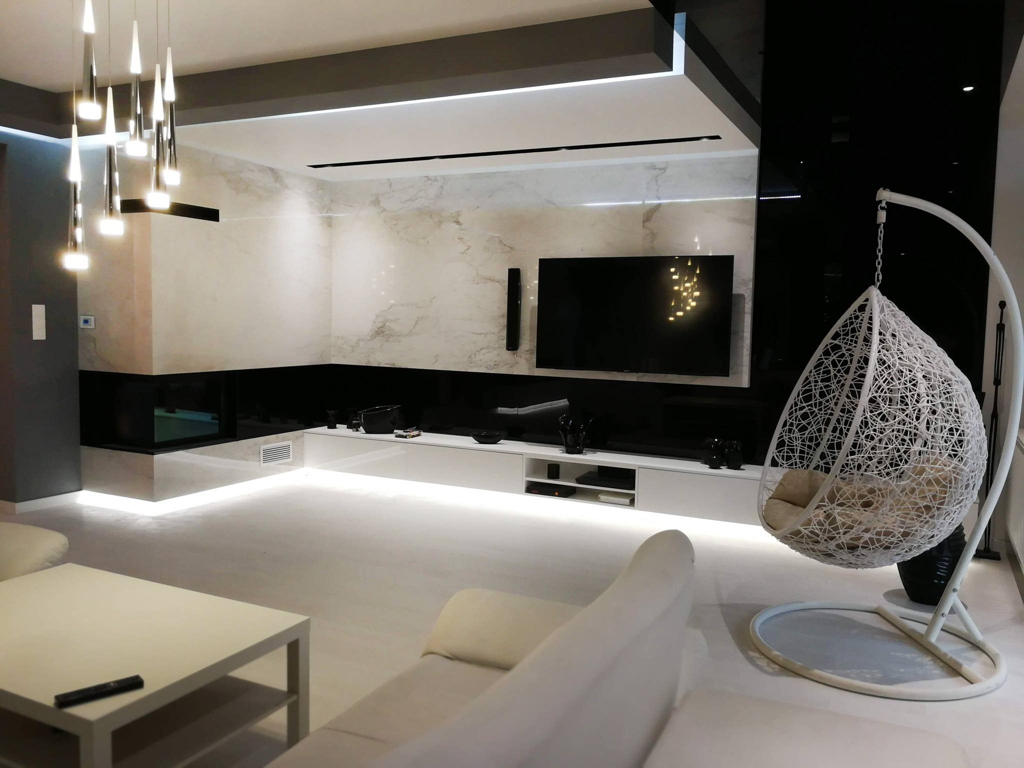 Nowoczesny salon z białymi meblami, marmurową ścianą, wiszącym fotelem typu kokon i designerską lampą wiszącą.