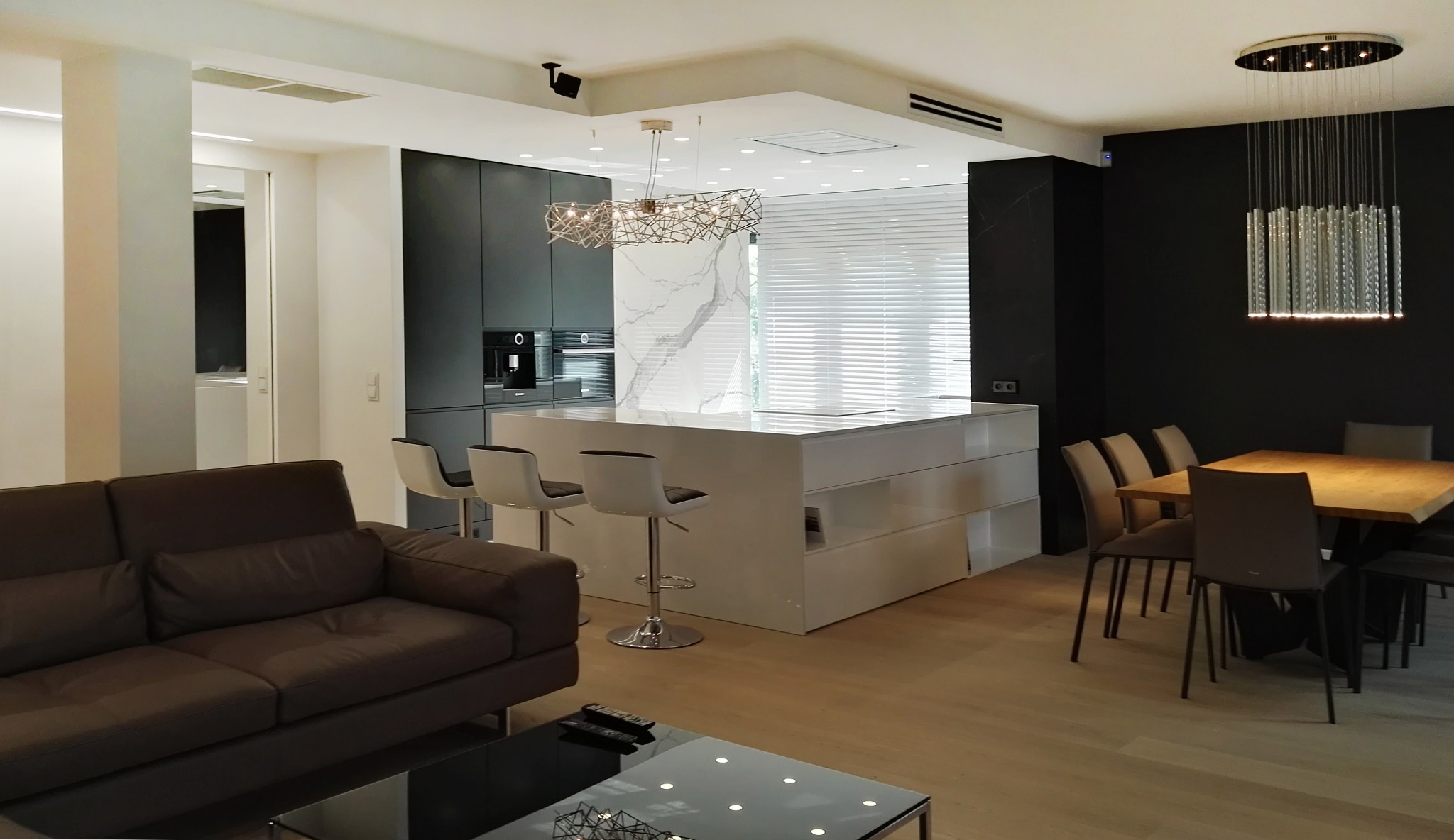 Nowoczesne wnętrze: salon z brązową skórzaną sofą, szklanym stolikiem kawowym, białą wyspą kuchenną z hokerami, czarną zabudową kuchenną, drewnianym stołem jadalnym i designerskim oświetleniem.