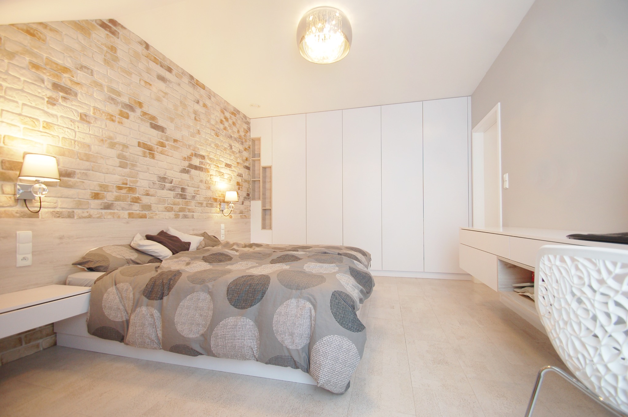 Jasna sypialnia z łóżkiem w szaro-białej pościeli w duże koła, ściana wyłożona cegłą w odcieniach beżu, białe szafy w zabudowie i lampy ścienne z kryształkami.
