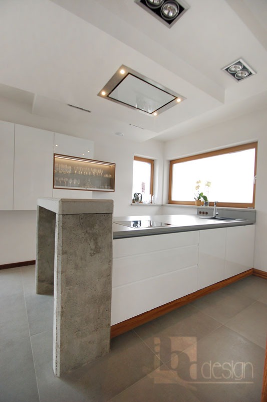 Nowoczesna kuchnia z wyspą o betonowych nogach, białe fronty szafek, drewniane wykończenia, okno z drewnianą ramą i oświetlenie sufitowe.