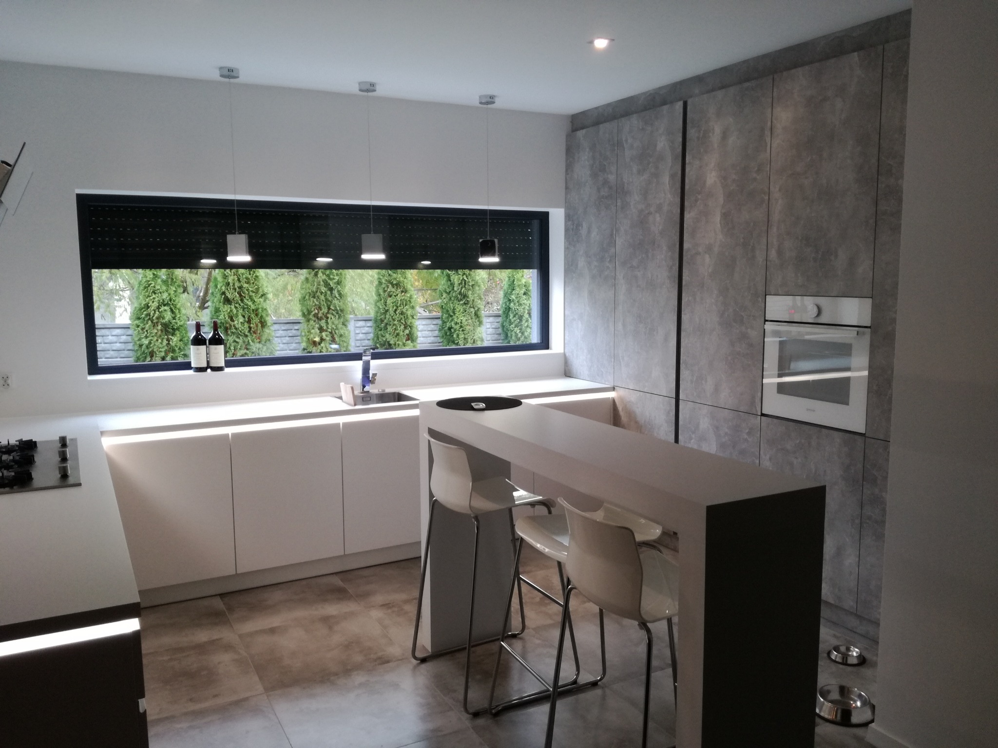 Nowoczesna kuchnia z wyspą, białymi szafkami, szarymi panelami imitującymi beton, oknem z widokiem na tuje i butelkami wina na parapecie, oraz miskami dla psa przy ścianie.