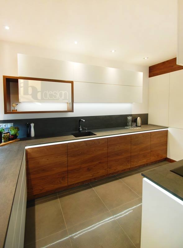 Nowoczesna kuchnia z drewnianymi szafkami dolnymi podświetlonymi LED, czarnym zlewem i blatem, białymi szafkami górnymi oraz półką z logo firmy projektowej.