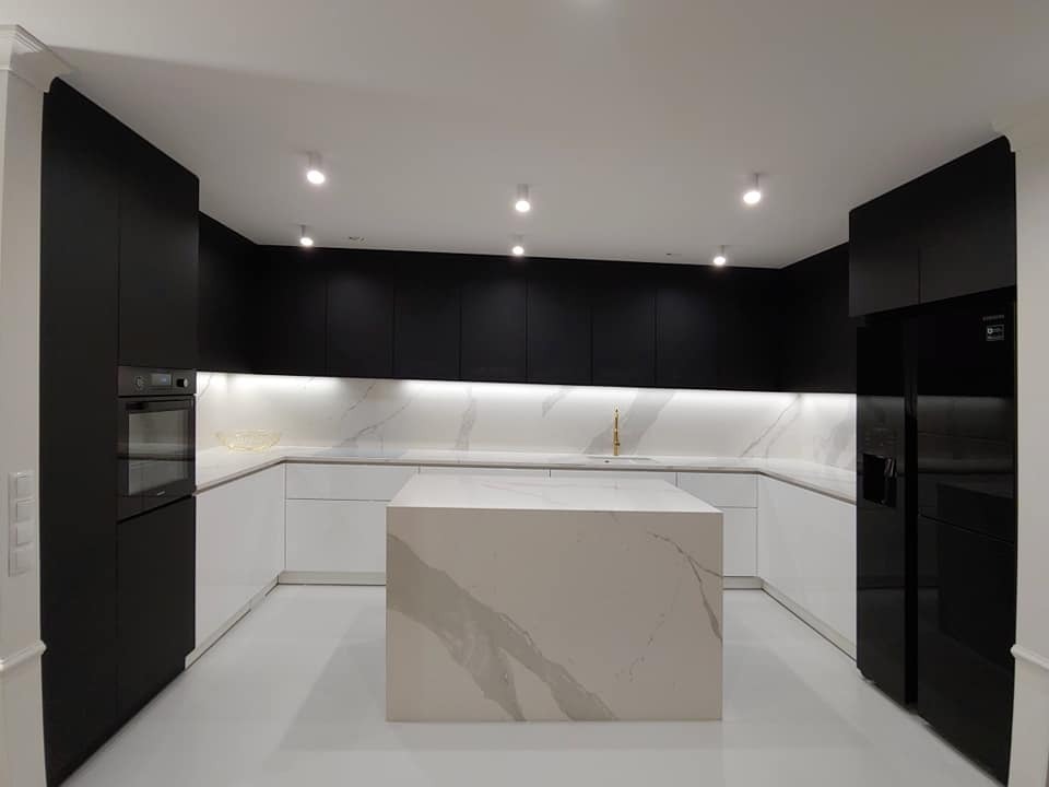 Minimalistyczna kuchnia z czarnymi szafkami górnymi i białymi dolnymi, wyspa z blatem imitującym marmur, złota bateria przy zlewozmywaku, oświetlenie punktowe.