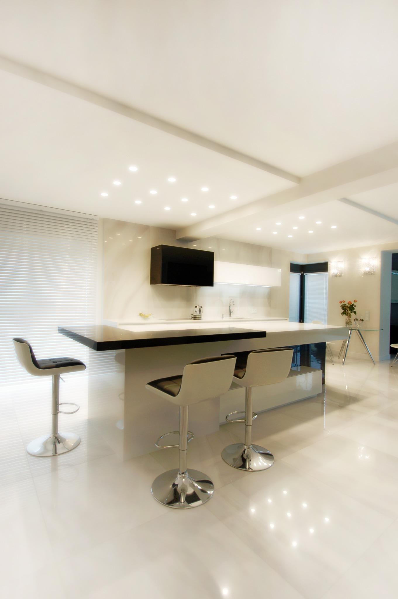 Nowoczesna kuchnia z wyspą, białe płytki podłogowe z połyskiem, czarny blat wyspy, chromowane hokery z białymi siedziskami, punktowe oświetlenie sufitowe.