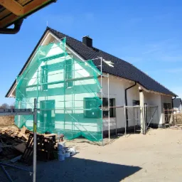 Dom jednorodzinny w trakcie ocieplania elewacji, widoczne rusztowania z siatką ochronną, stos desek i wiader z farbą, surowy teren budowy i zaparkowany biały samochód osobowy.