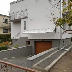 Dwupiętrowy dom z białymi ścianami, balkonem z ozdobną balustradą, szarą elewacją parteru i drewnianą bramą garażową pod zadaszeniem z poliwęglanu.