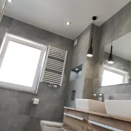 Nowoczesna łazienka z szarymi płytkami, drewnianą szafką pod umywalką, białymi umywalkami nablatowymi i grzejnikiem drabinkowym. Widoczne czarne lampy wiszące i okno z białym profilem.