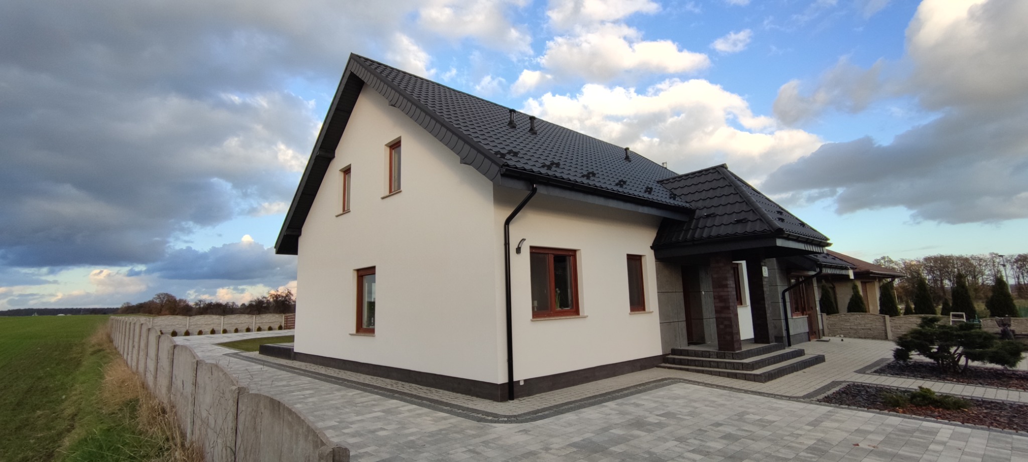 Nowoczesny dom murowany z białą elewacją, ciemną dachówką i drewnianymi oknami, otoczony brukowanym podjazdem i betonowym ogrodzeniem, na tle pochmurnego nieba.