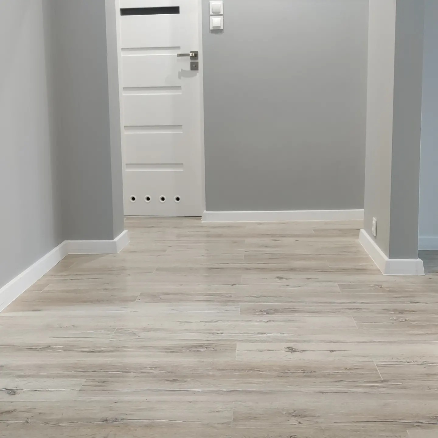 Korytarz z panelami podłogowymi imitującymi drewno, szare ściany z białymi listwami przypodłogowymi, białe drzwi z poziomymi frezowaniami i czterema okrągłymi otworami wentylacyjnymi u dołu.