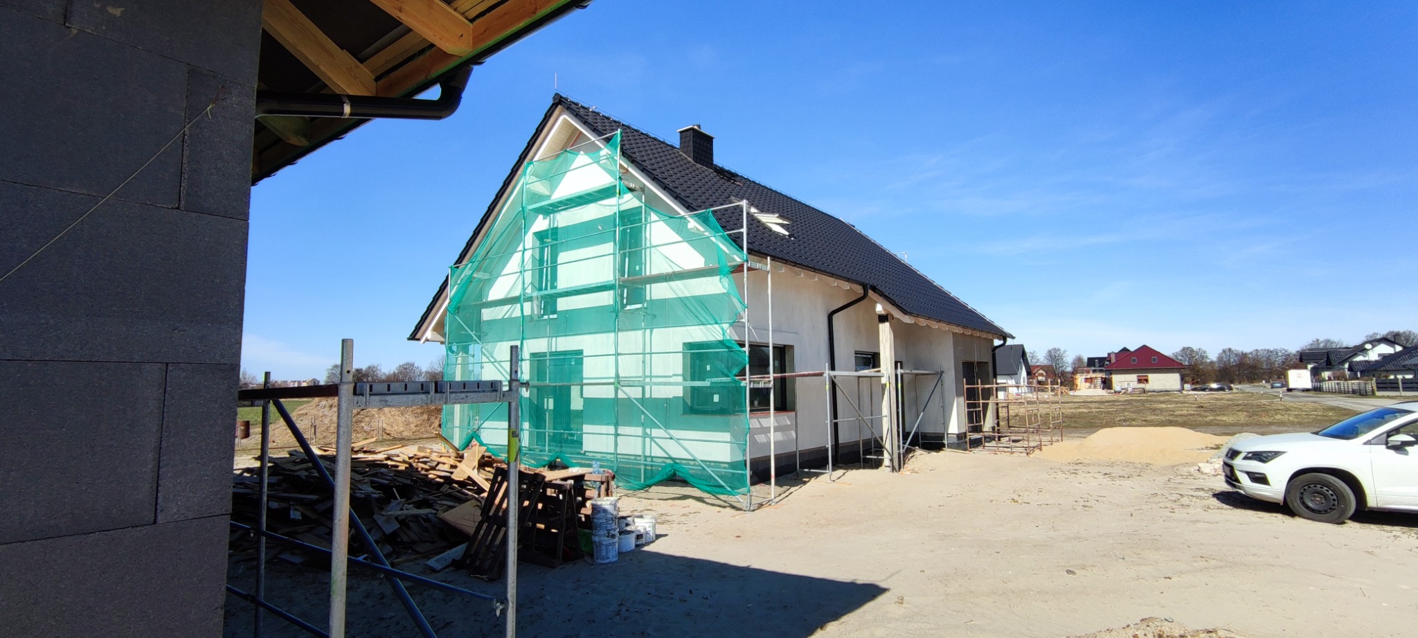 Dom jednorodzinny w trakcie ocieplania elewacji, widoczne rusztowania z siatką ochronną, stos desek i wiader z farbą, surowy teren budowy i zaparkowany biały samochód osobowy.