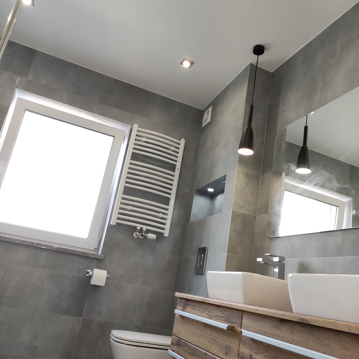 Nowoczesna łazienka z szarymi płytkami, drewnianą szafką pod umywalką, białymi umywalkami nablatowymi i grzejnikiem drabinkowym. Widoczne czarne lampy wiszące i okno z białym profilem.