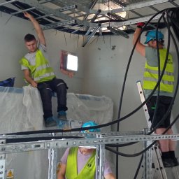 SERHII DOMIN DOMINWEST ENERGI - Trzech elektryków w kamizelkach odblaskowych montuje koryta kablowe pod sufitem w pomieszczeniu technicznym. Jeden siedzi na osłoniętym folią urządzeniu, drugi stoi na drabinie, a trzeci manipuluje...