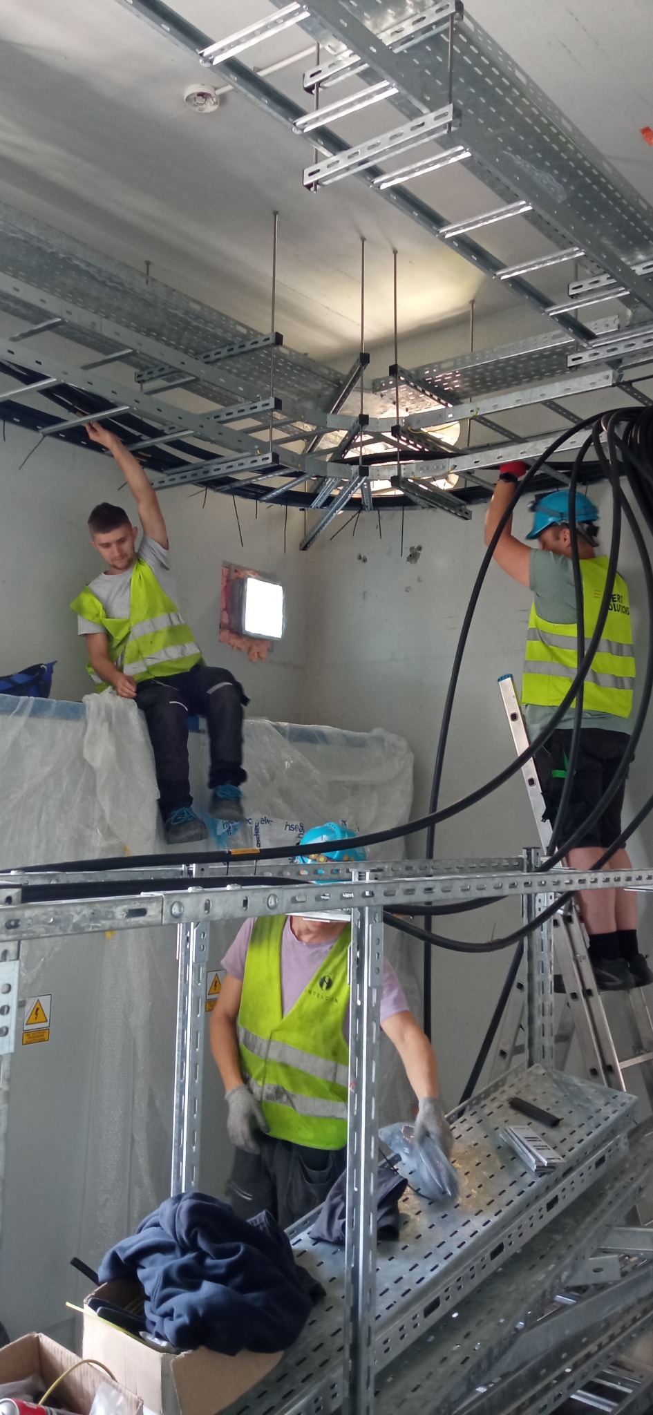 Trzech elektryków w kamizelkach odblaskowych montuje koryta kablowe pod sufitem w pomieszczeniu technicznym. Jeden siedzi na osłoniętym folią urządzeniu, drugi stoi na drabinie, a trzeci manipuluje...