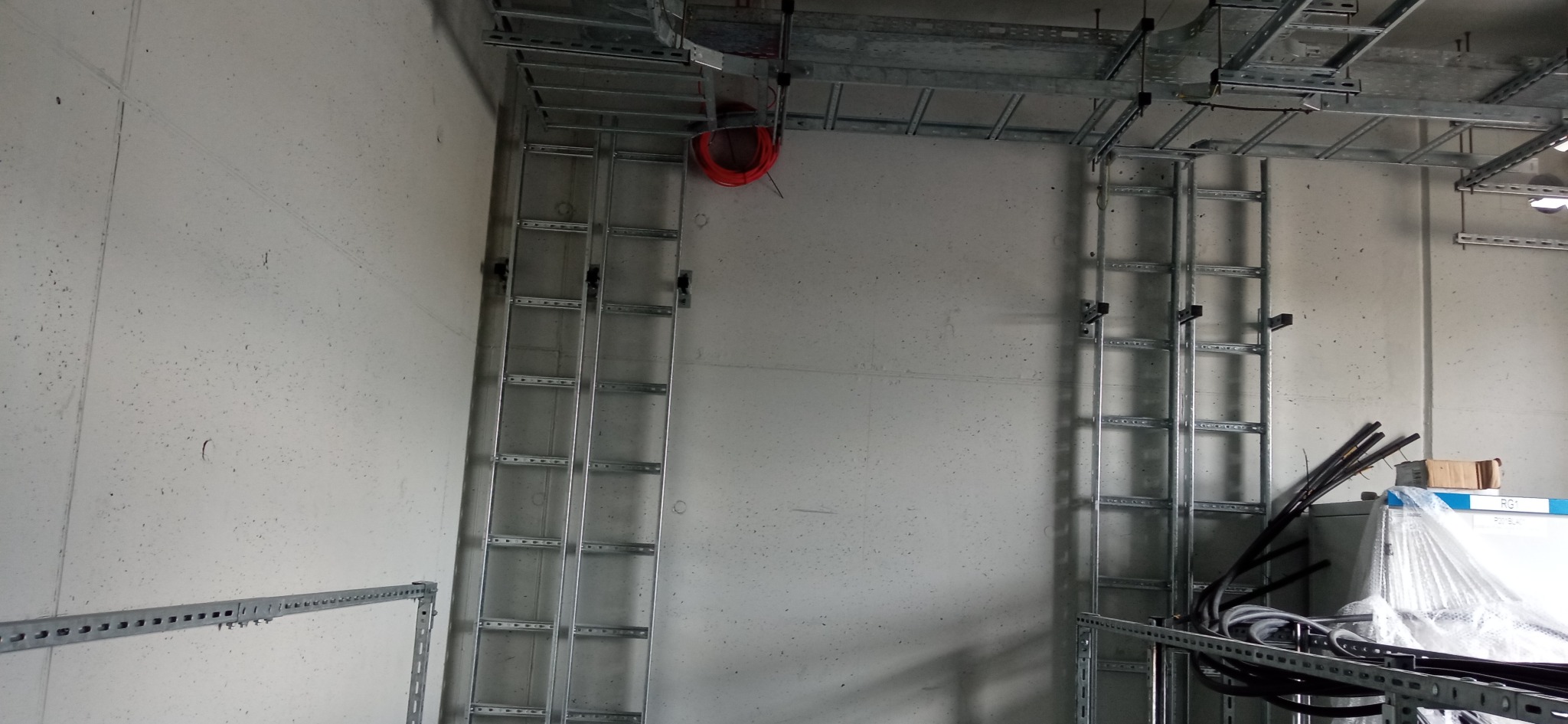 Metalowe korytka kablowe zamontowane na betonowej ścianie w hali przemysłowej, z widocznymi kablami i elementami instalacji pod sufitem.