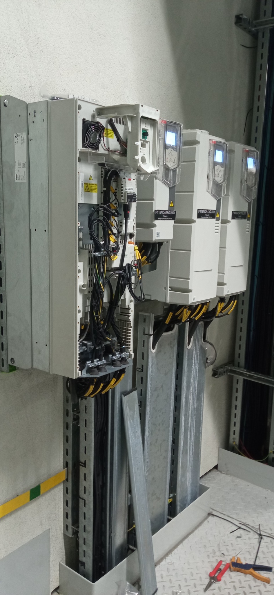 Trzy przemienniki częstotliwości ABB zamontowane na ścianie, jeden z otwartą obudową ukazującą wewnętrzne okablowanie i wentylator, z widocznymi przewodami w żółtej osłonie i narzędziami na podłodze.