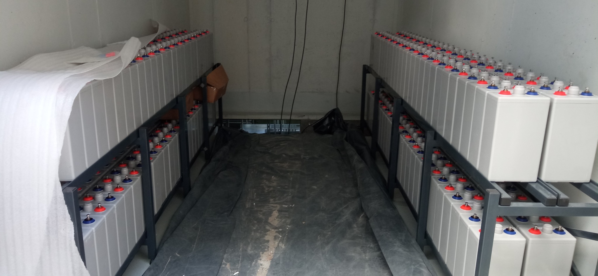 Rzędy akumulatorów przemysłowych z czerwonymi i niebieskimi korkami na metalowych regałach w pomieszczeniu technicznym, zabezpieczone folią ochronną.