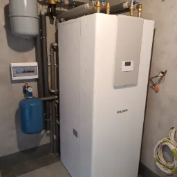 Nowoczesny biały piec gazowy marki Stiebel Eltron z szarymi elementami, podłączony do instalacji z izolowanymi rurami, zbiornikiem wyrównawczym i rozdzielnicą elektryczną na tle szarych płytek.