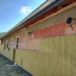 Budynek w trakcie ocieplania: ściany z czerwonej cegły częściowo pokryte żółtą wełną mineralną, widoczne elementy wentylacji i rynna, na zewnątrz zielony teren.