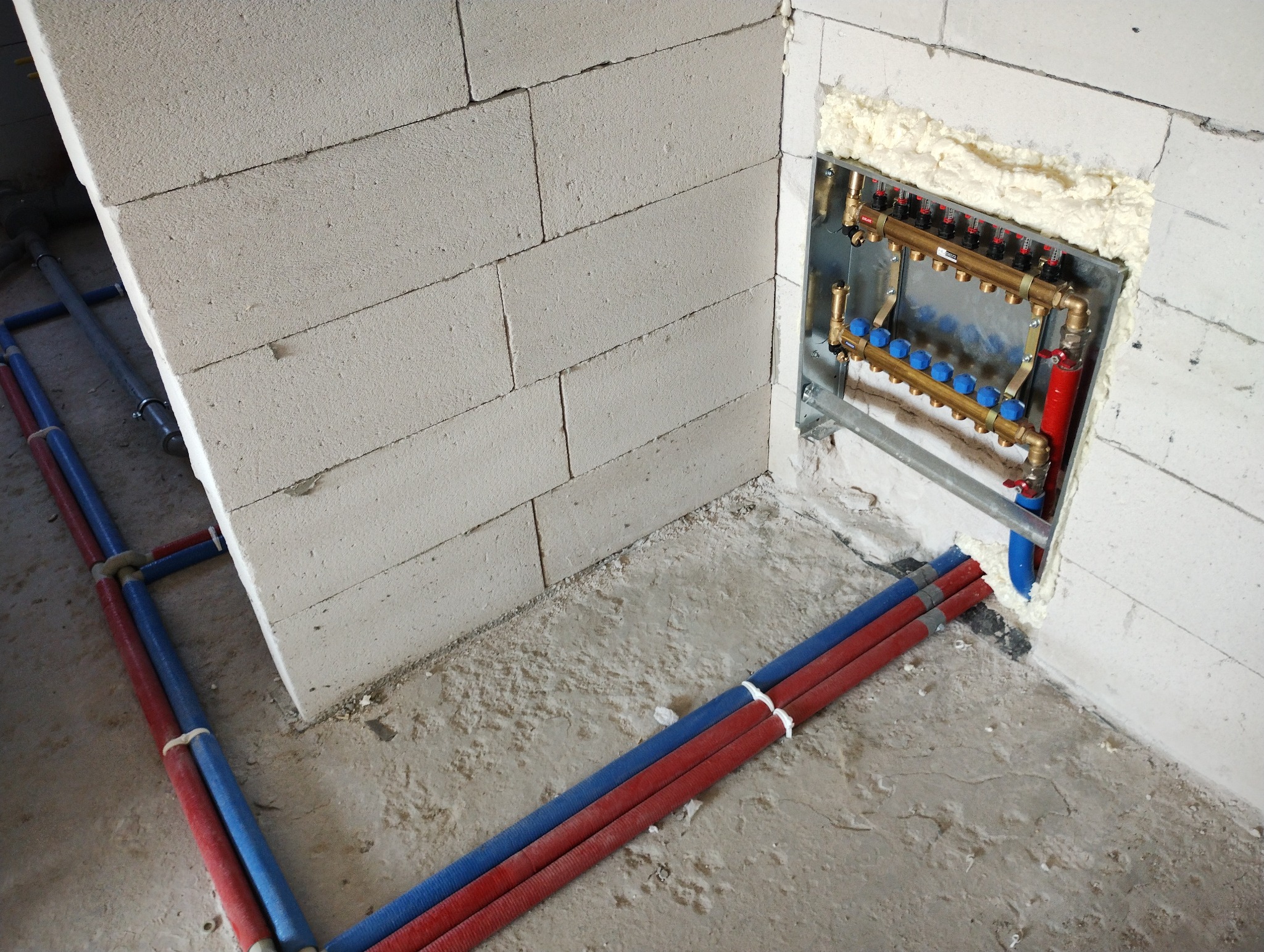 Rozdzielacz centralnego ogrzewania zamontowany w ścianie z betonu komórkowego, z widocznymi rurami doprowadzającymi w kolorze czerwonym i niebieskim, zabezpieczonymi pianką montażową.