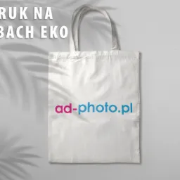 Biała torba bawełniana z nadrukowanym adresem strony internetowej 'ad-photo.pl' w kolorach magenta i błękitu, leżąca na szarym tle z cieniem liści.