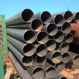 Stos okrągłych, stalowych rur na zielonym, metalowym stojaku, widoczne ślady rdzy i zarysowania, błękitne niebo w tle.