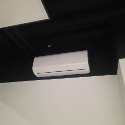 Biały klimatyzator marki Rotenso zamontowany na ścianie w pomieszczeniu z czarnym sufitem i białą ścianą, widok z dołu.