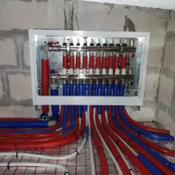 Rozdzielacz ogrzewania podłogowego w skrzynce, z czerwonymi i niebieskimi rurami na siatce, surowa ściana w tle.