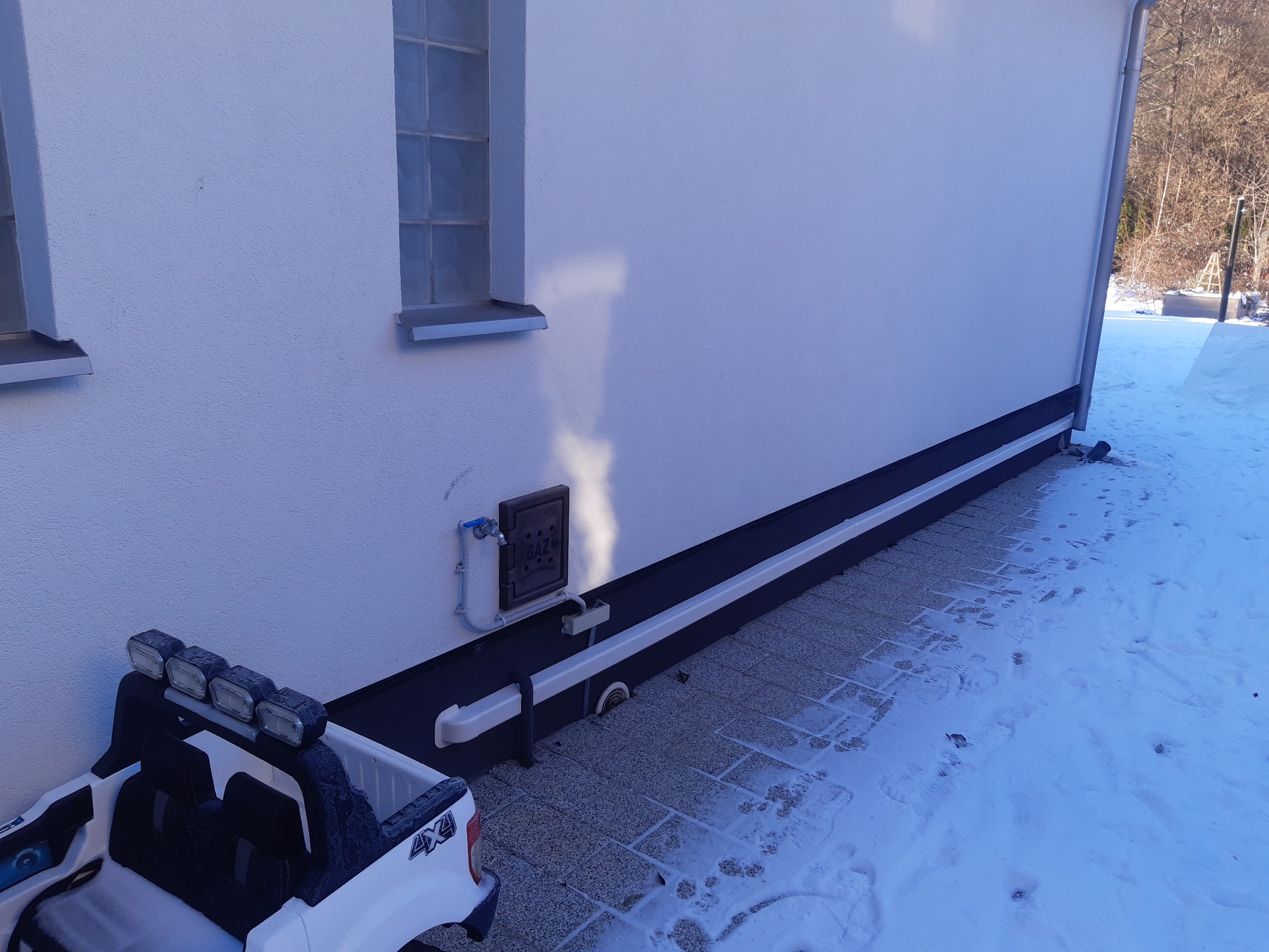 Biała ściana budynku z oknem i skrzynką gazową, na pierwszym planie zabawkowy samochód terenowy z oświetleniem dachowym.