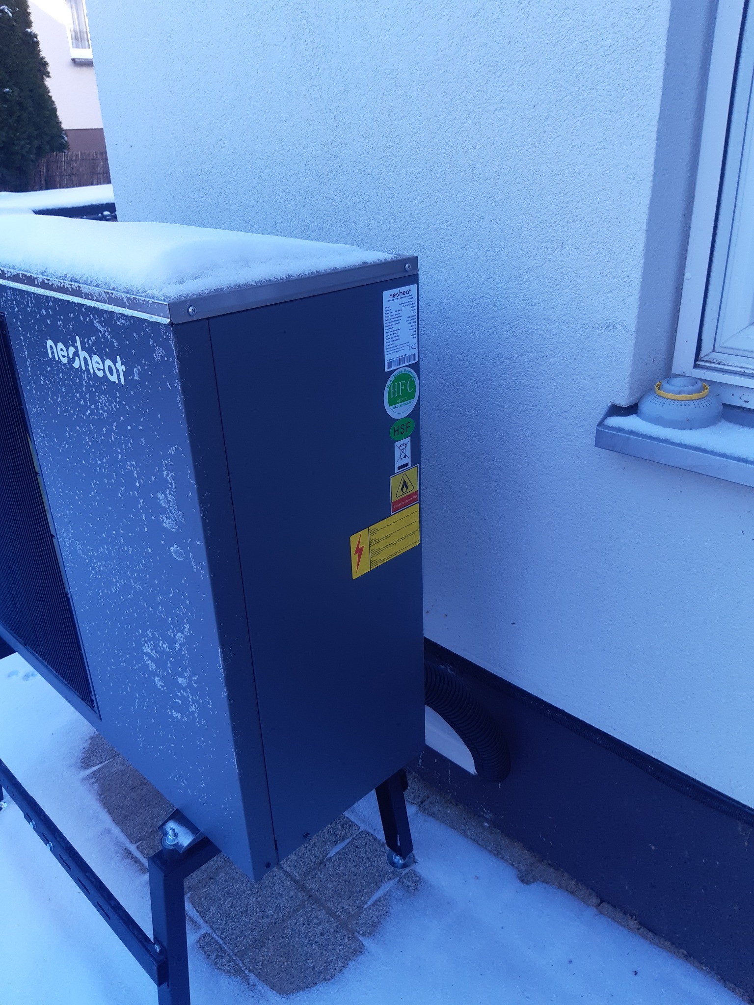 Zewnętrzna jednostka pompy ciepła marki Nesheat, lekko przysypana śniegiem, zamontowana na metalowej konstrukcji przy ścianie budynku z widocznym oknem i fragmentem przewodu odprowadzającego...